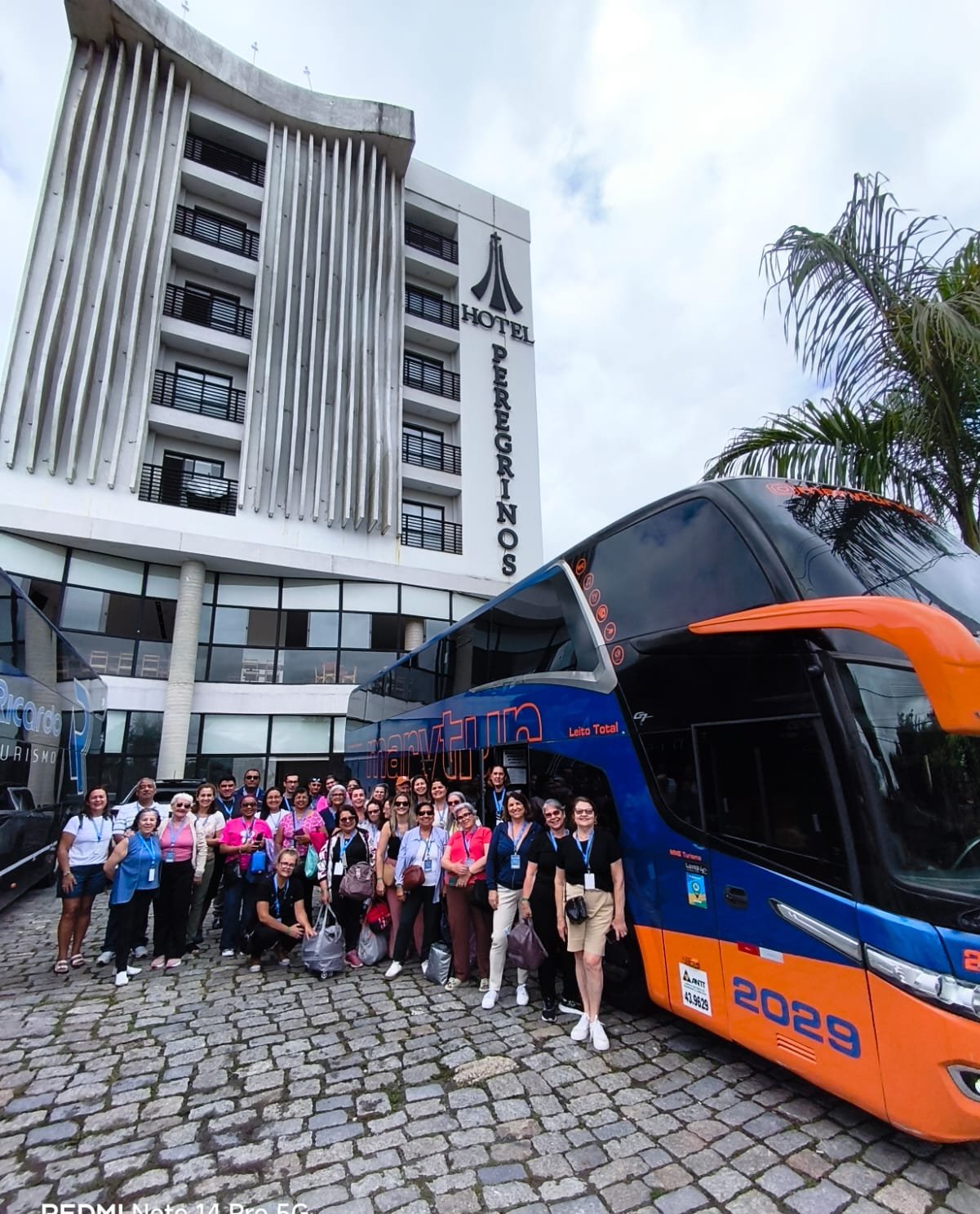 Grupo em frente ao ônibus azul e laranja da agência, pátio com edifício ao fundo