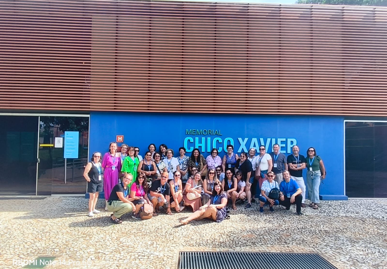 Grupo grande reunido em frente à estátua de Chico Xavier com céu azul ao fundo