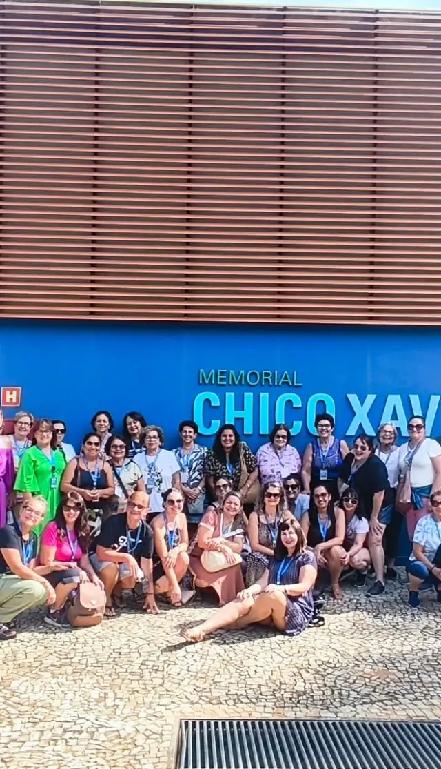 Grupo grande reunido em frente à estátua de Chico Xavier com céu azul ao fundo